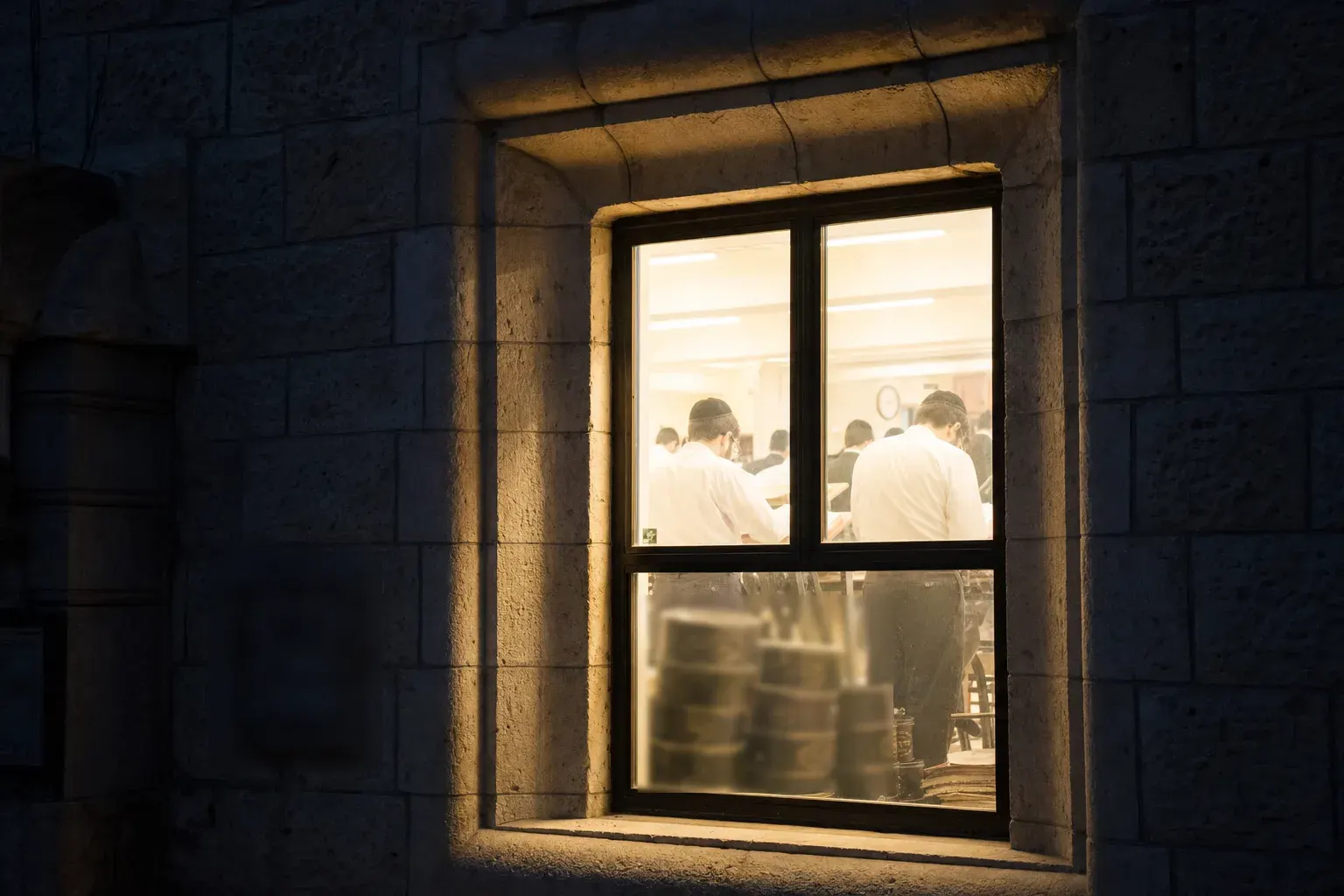 One lit window of a Jerusalem-stone building at night, learners visible from behind through the glass