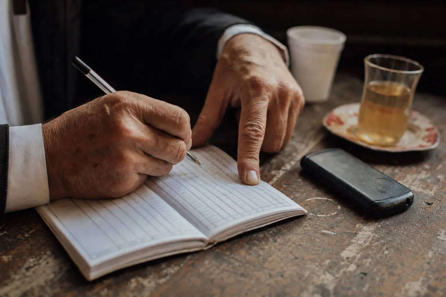 Weathered hands over a small notebook, a phone and a tea glass beside it