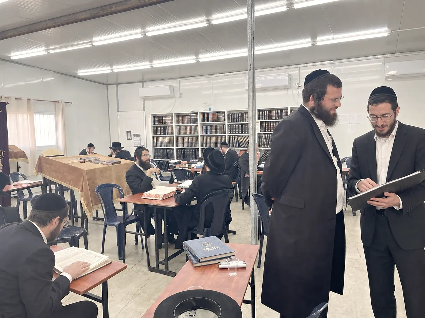 A visitor standing inside a kollel in conversation while avreichim learn at the tables around him