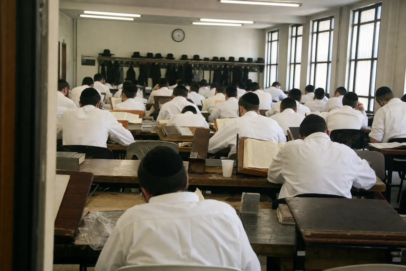 A full beis medrash seen from the doorway, every yungerman bent over his Gemara, hats along the rail